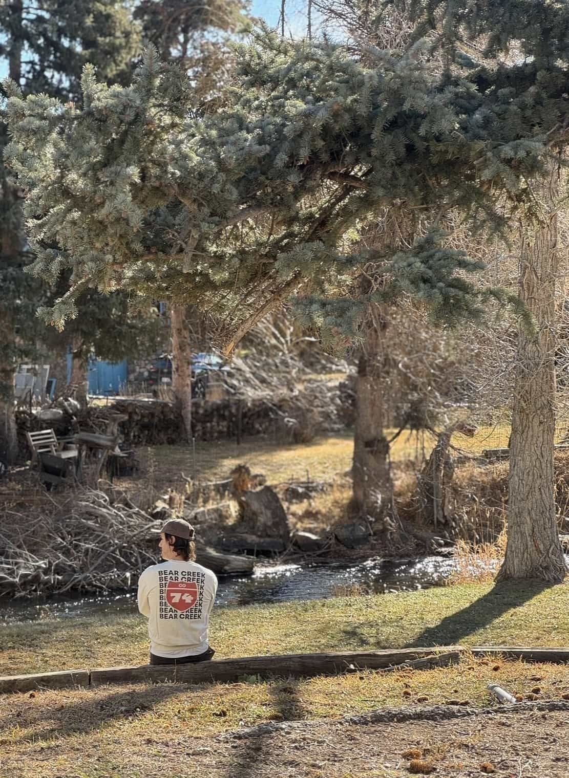 A runner in a CO74 Bear Creek t-shirt sitting beside Bear Creek under a blue spruce.