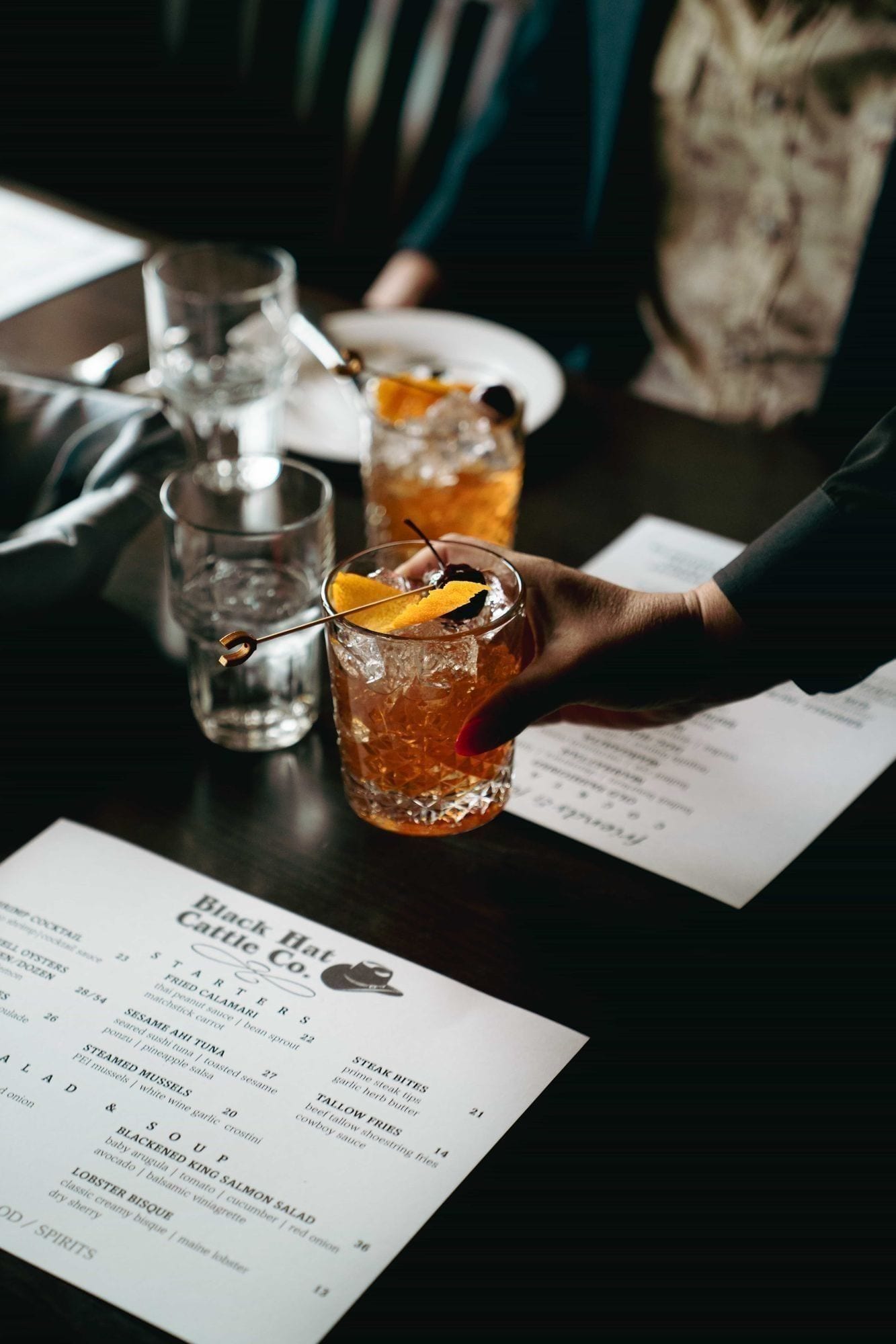 A round of drinks set out on a wooden table at Black Hat Cattle Co — beers, cocktails, and a pair of shot glasses sweating in the late-afternoon light.