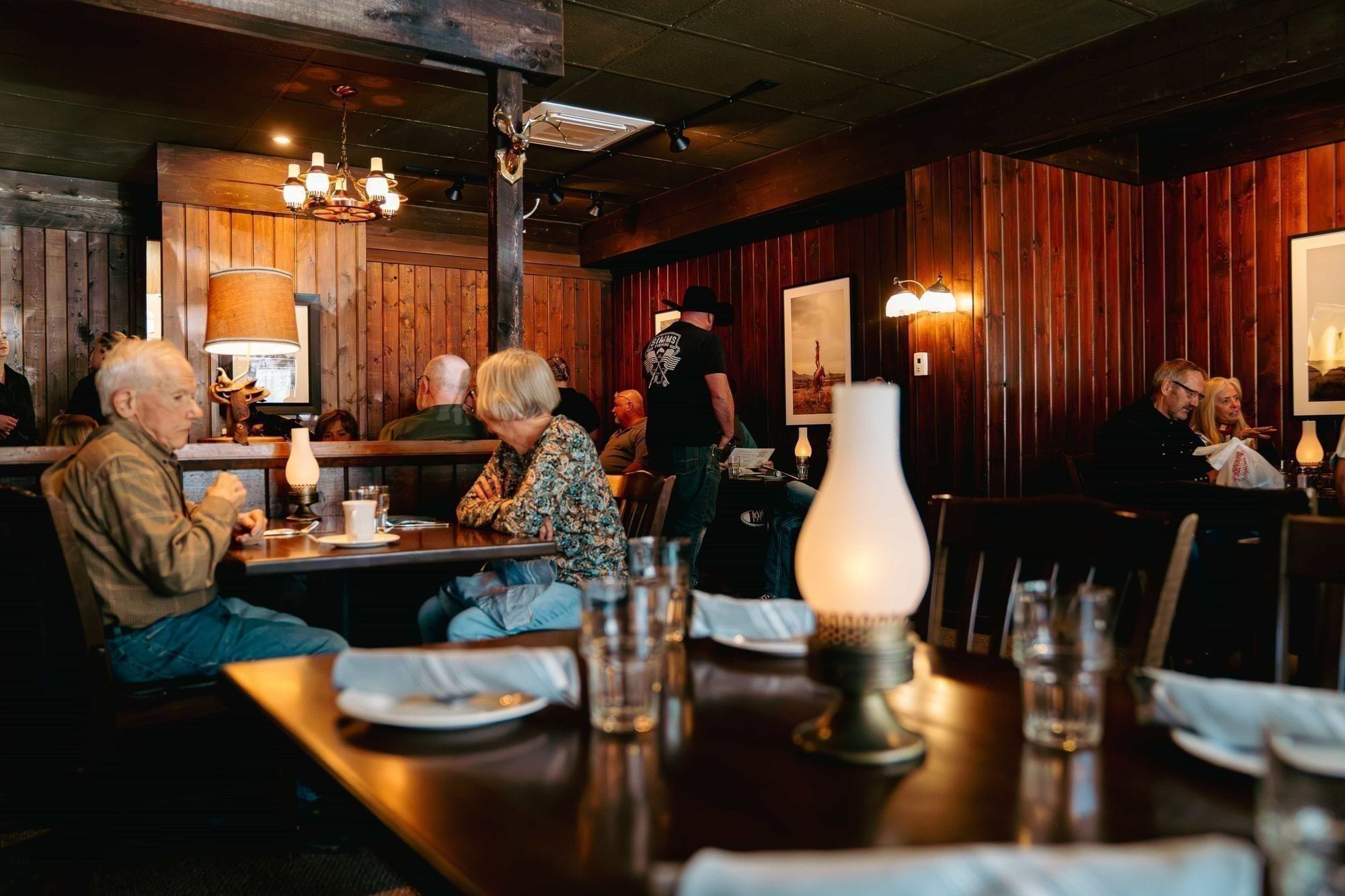 A server carrying a tray of plates through the dining room at Black Hat Cattle Co, the room full of guests at long communal tables.