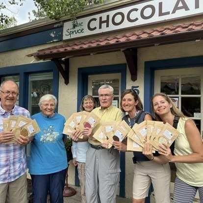 Customers gathered outside Blue Spruce Chocolates on a sunny Kittredge afternoon, boxes of bonbons in hand and the storefront sign visible behind them.