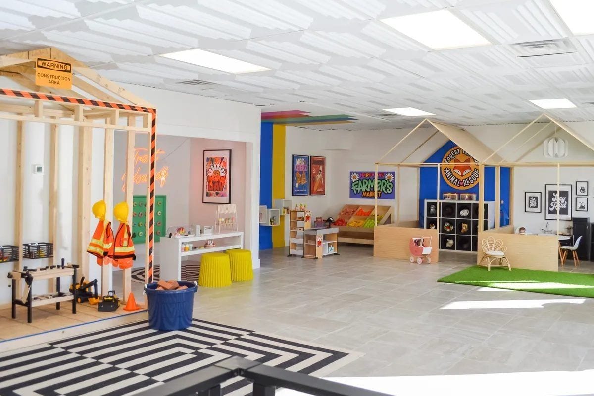 Inside Happy Day — bright, plant-filled main room with kid-sized tables, art on the walls, and a reading nook in the back corner.