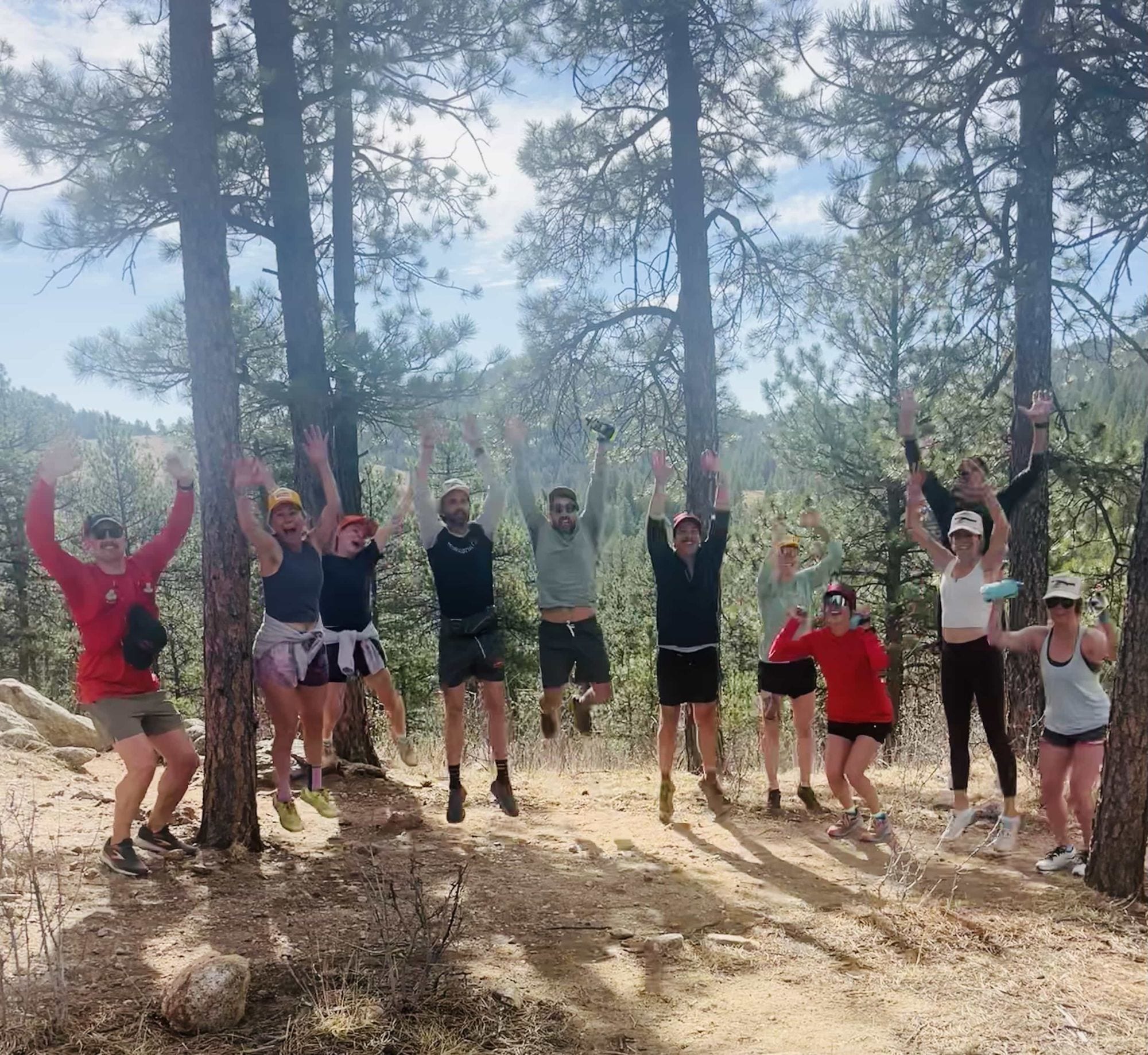Kittredge Run Club members leaping into the air mid-celebration on a sunlit trail through ponderosa pines.