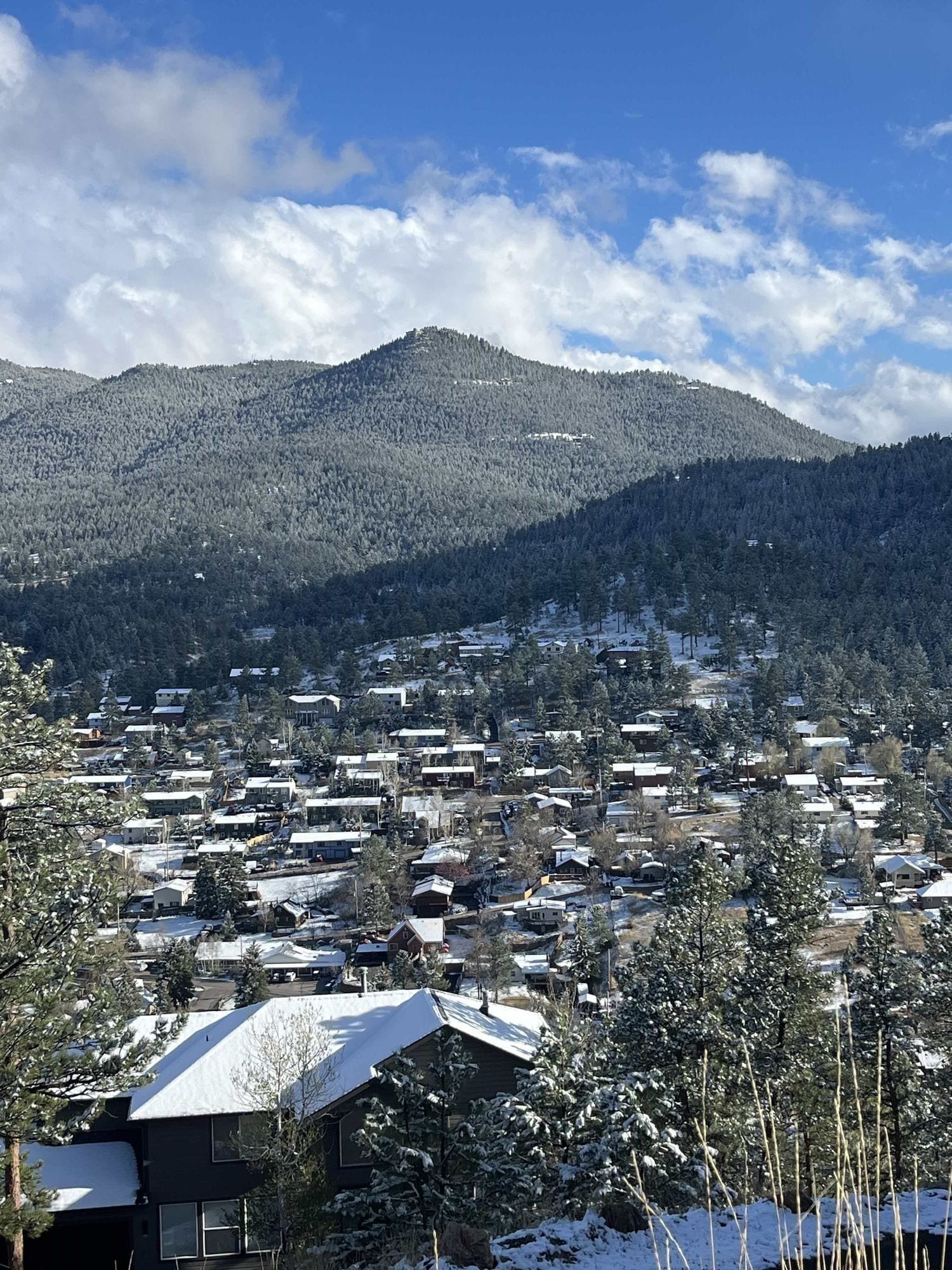 Snow-dusted rooftops of Kittredge homes with a tall forested mountain rising behind them under a blue sky.