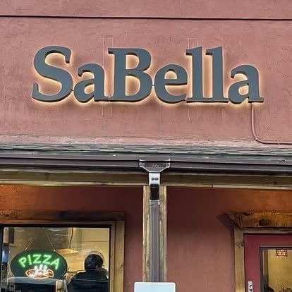 The hand-painted Sabella Pizzeria sign on the side of the building — bold serif lettering, a sliver of the open dining room visible through the window.